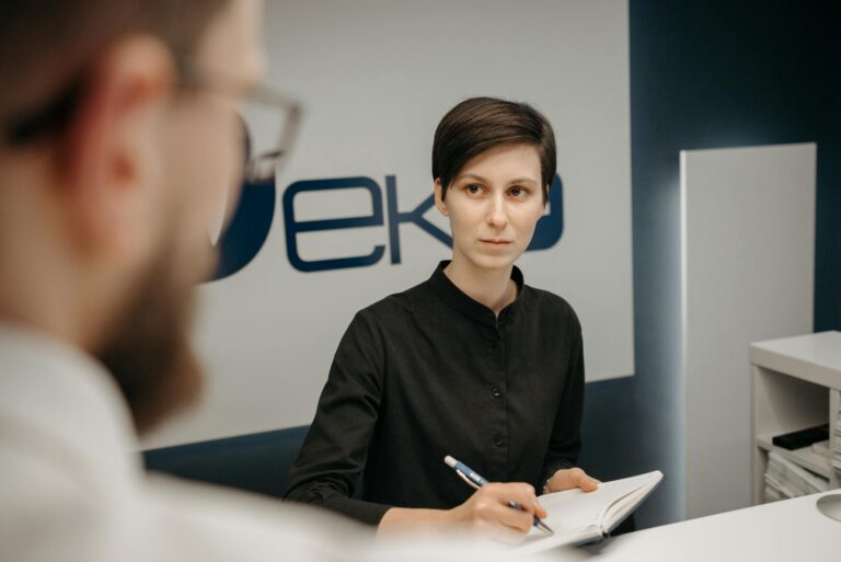 A receptionist at a clinic desk writing notes during a consultation.